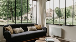 Ein modernes Wohnzimmer mit einem dunklen Sofa, zwei Kissen und einem runden Couchtisch. Große Fenster bieten Blick auf einen Garten und ein Gebäude im Hintergrund.