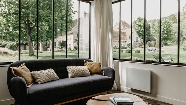 Een moderne woonkamer met een donkere bank, twee kussens en een ronde houten salontafel. Grote ramen bieden uitzicht op een tuin en een gebouw op de achtergrond.