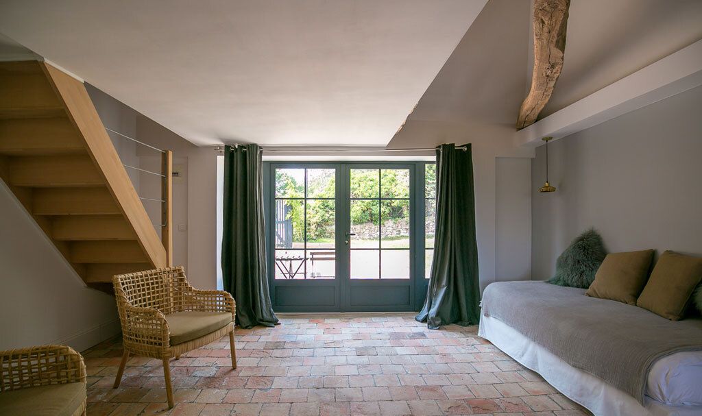 A room with a bed, a wicker chair, a staircase, and a large window with curtains.
