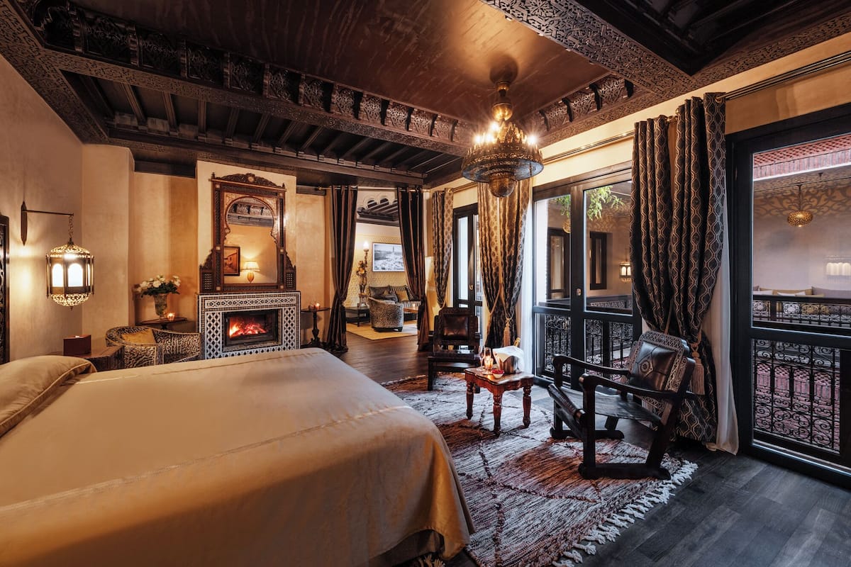 Un dormitorio lujoso con una cama grande, chimenea, techo ornamentado y un balcón con cortinas estampadas.