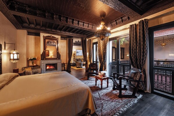 A luxurious bedroom with a large bed, a fireplace, ornate ceiling, and a balcony with patterned curtains.