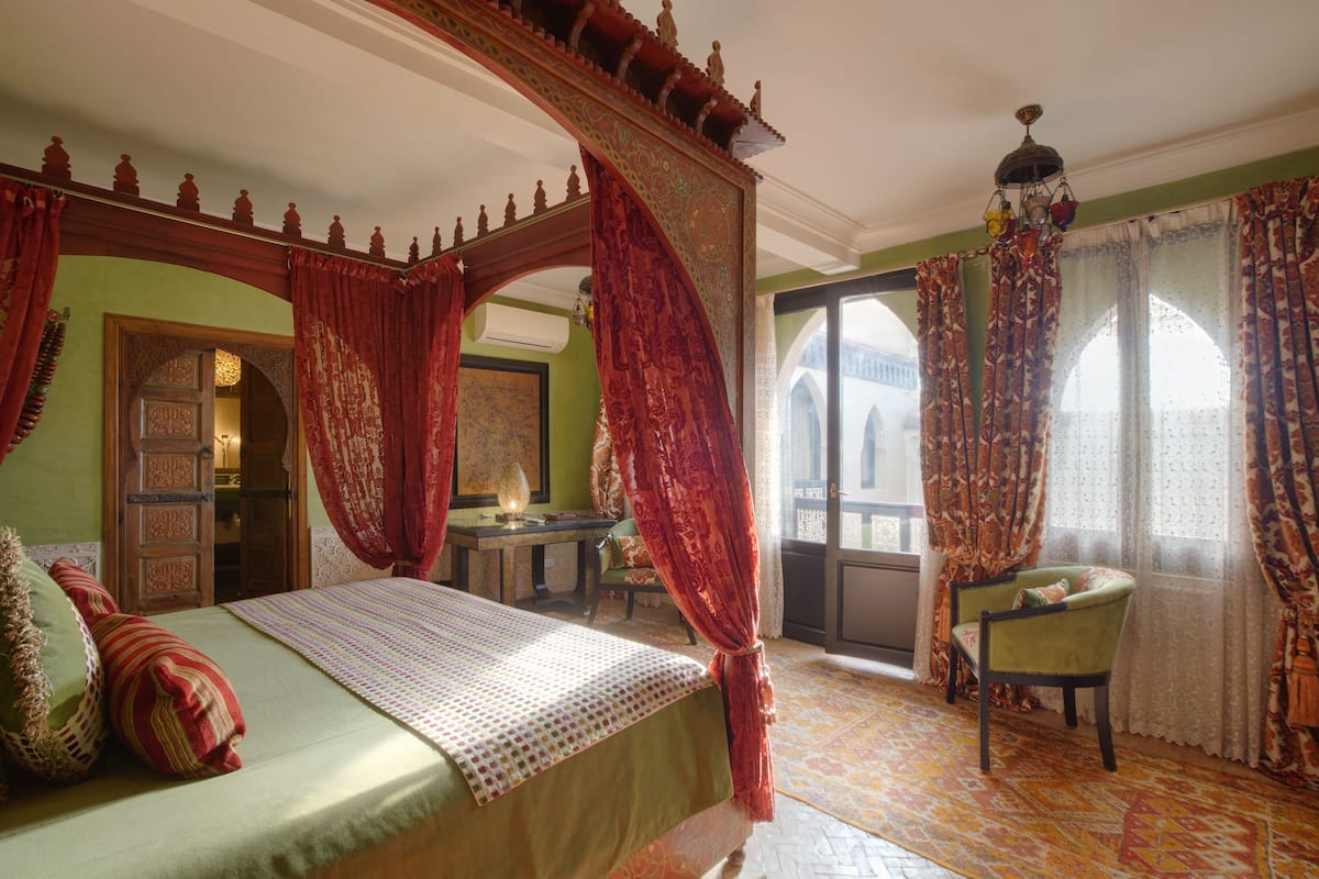 Una cama con dosel adornada con cortinas rojas, un sillón tapizado en verde y un suelo con diseño.