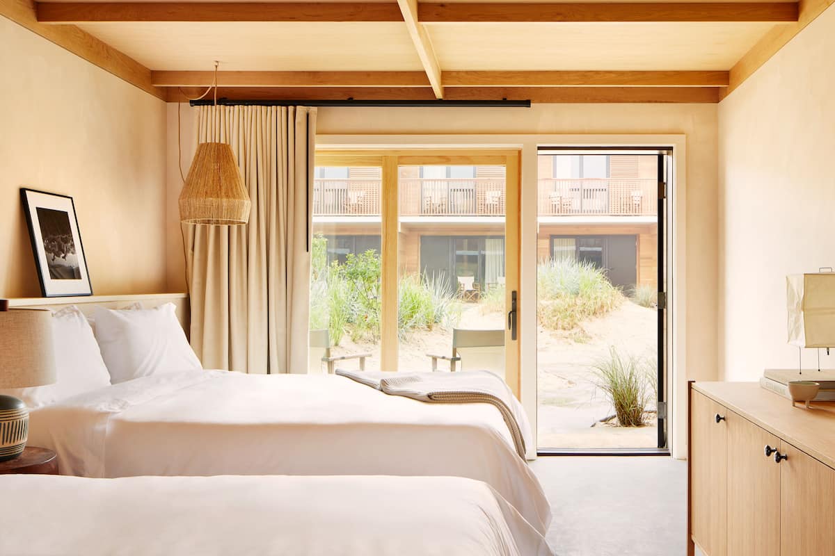 A hotel room with a large bed, a bedside table, a lamp, a framed picture, a hanging lamp, and a sliding glass door leading to an outdoor area with a view of a building and greenery.