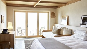 Habitación de hotel con un amplio ventanal corredero que da a una zona de estar exterior, cama con ropa de cama blanca, escritorio con lámpara y una lámpara colgante.