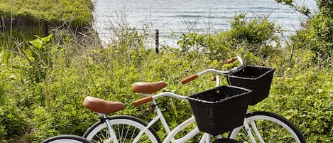 A white bicycle with a wicker basket and brown seat parked on a paved path by the water.
