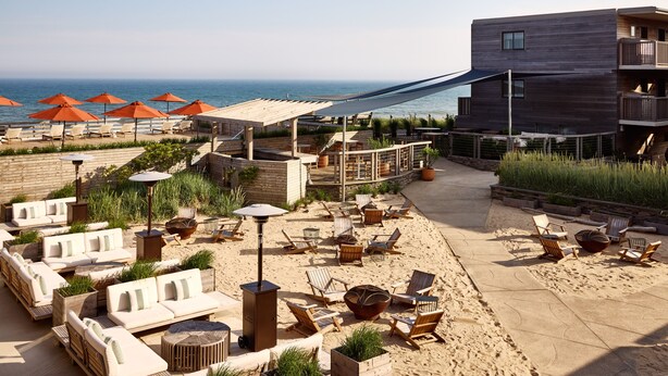 Un patio frente a la playa con sillones, sombrillas y un edificio de madera con balcón con vista al océano.