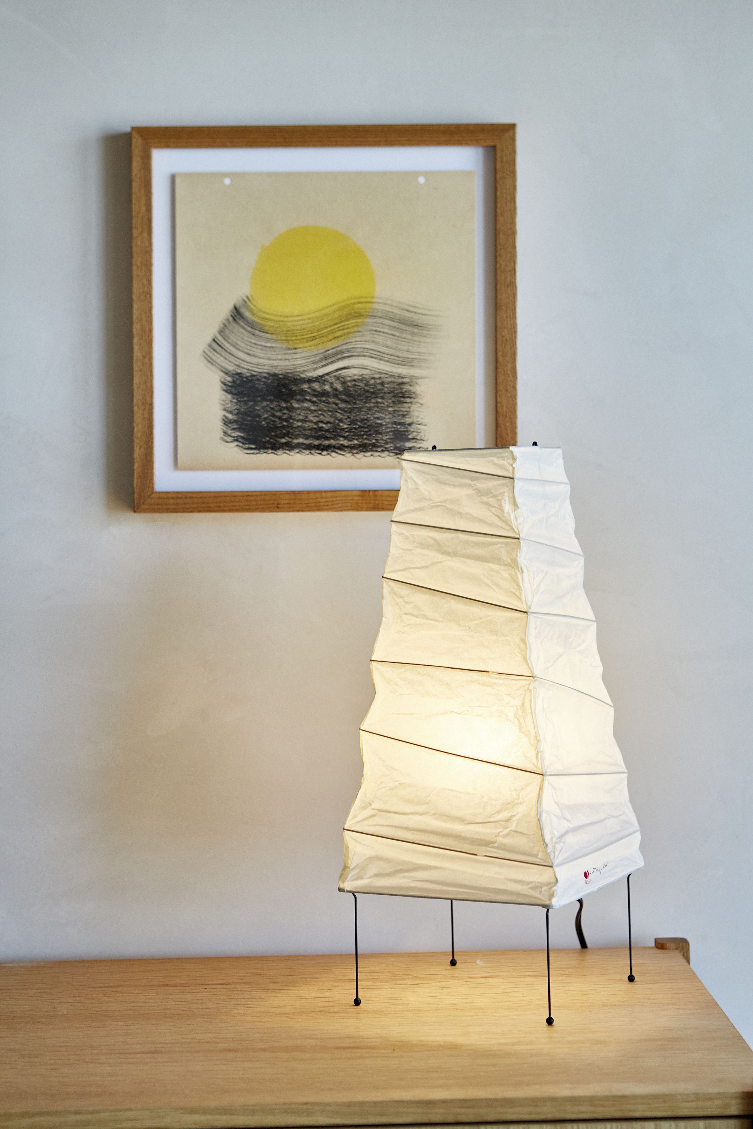 Una obra de arte enmarcadosi con un sol y formas abstractas en una pared, una silla blanca doblable con un logo en el respaldo, y una mesa de madera al frente.