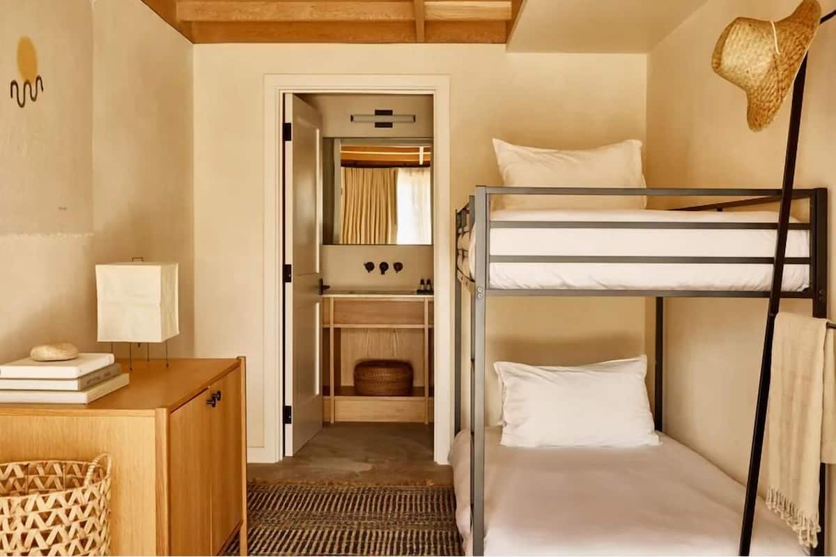 A compact hotel room with a bunk bed, a wooden cabinet, a desk with a lamp, and a wardrobe with a mirror.