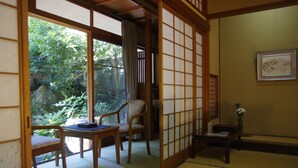 Una habitación tradicional japonesa con suelo de tatami, una mesa baja con cojín, una silla y una puerta corredera que da a un jardín.