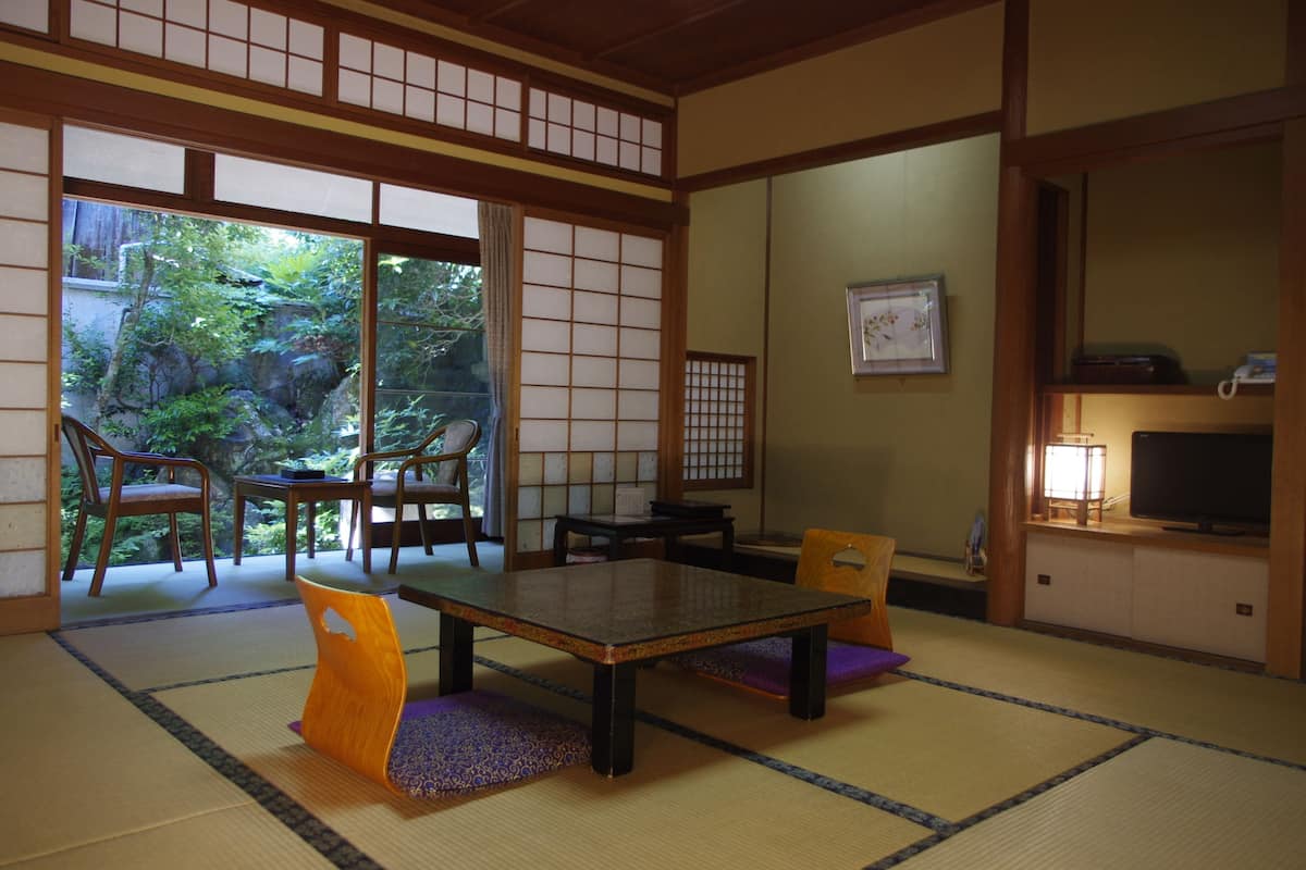 A traditional Japanese room with tatami flooring, a low table, chairs, and sliding doors leading to a garden.