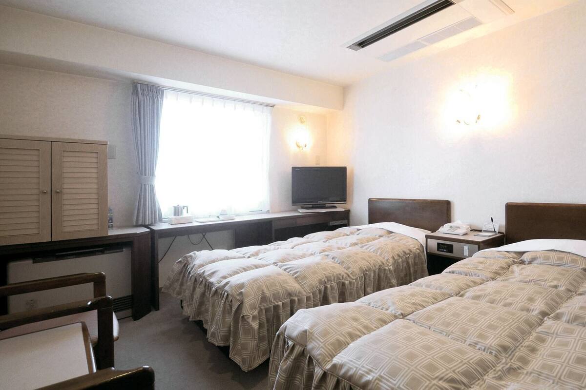 A hotel room with two beds, a desk with a television, a wardrobe, and a window with curtains.