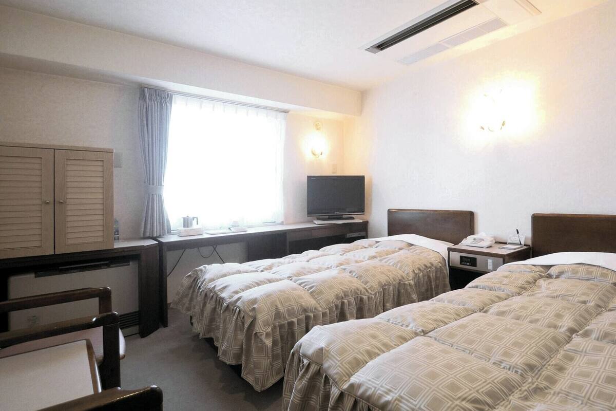 A hotel room with two beds, a desk with a television, a wardrobe, and a window with curtains.