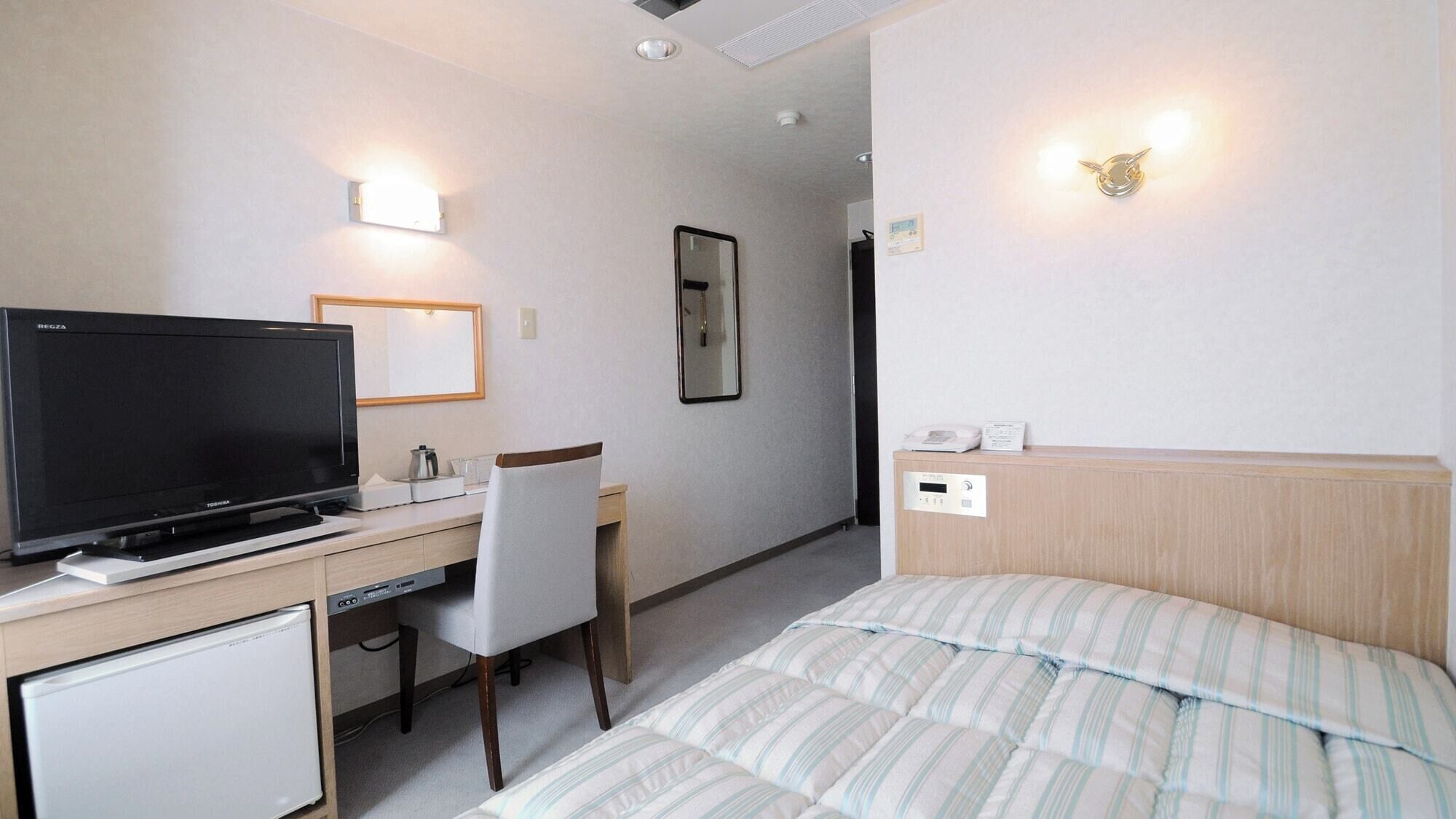 A hotel room with a bed, a desk with a television, a chair, a bedside table, and a wall-mounted light fixture.