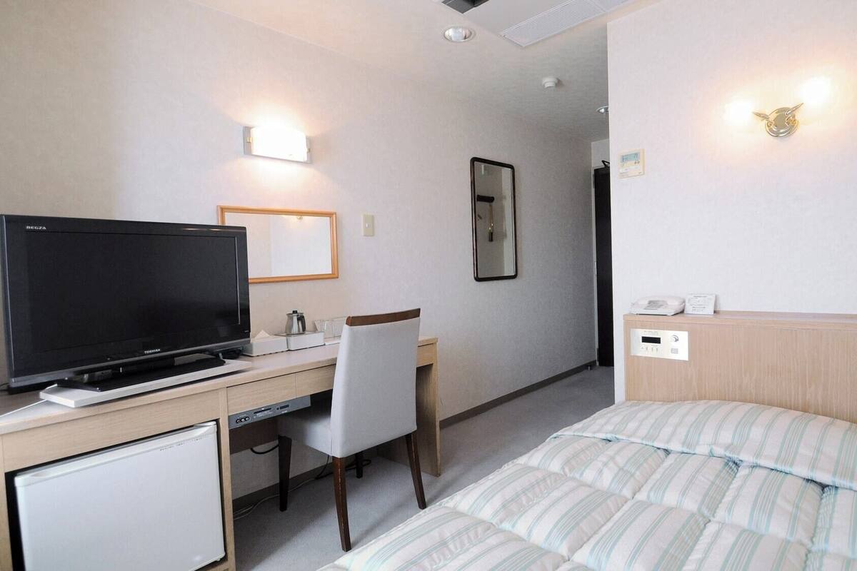 A hotel room with a bed, a desk with a television, a chair, a bedside table, and a wall-mounted light fixture.