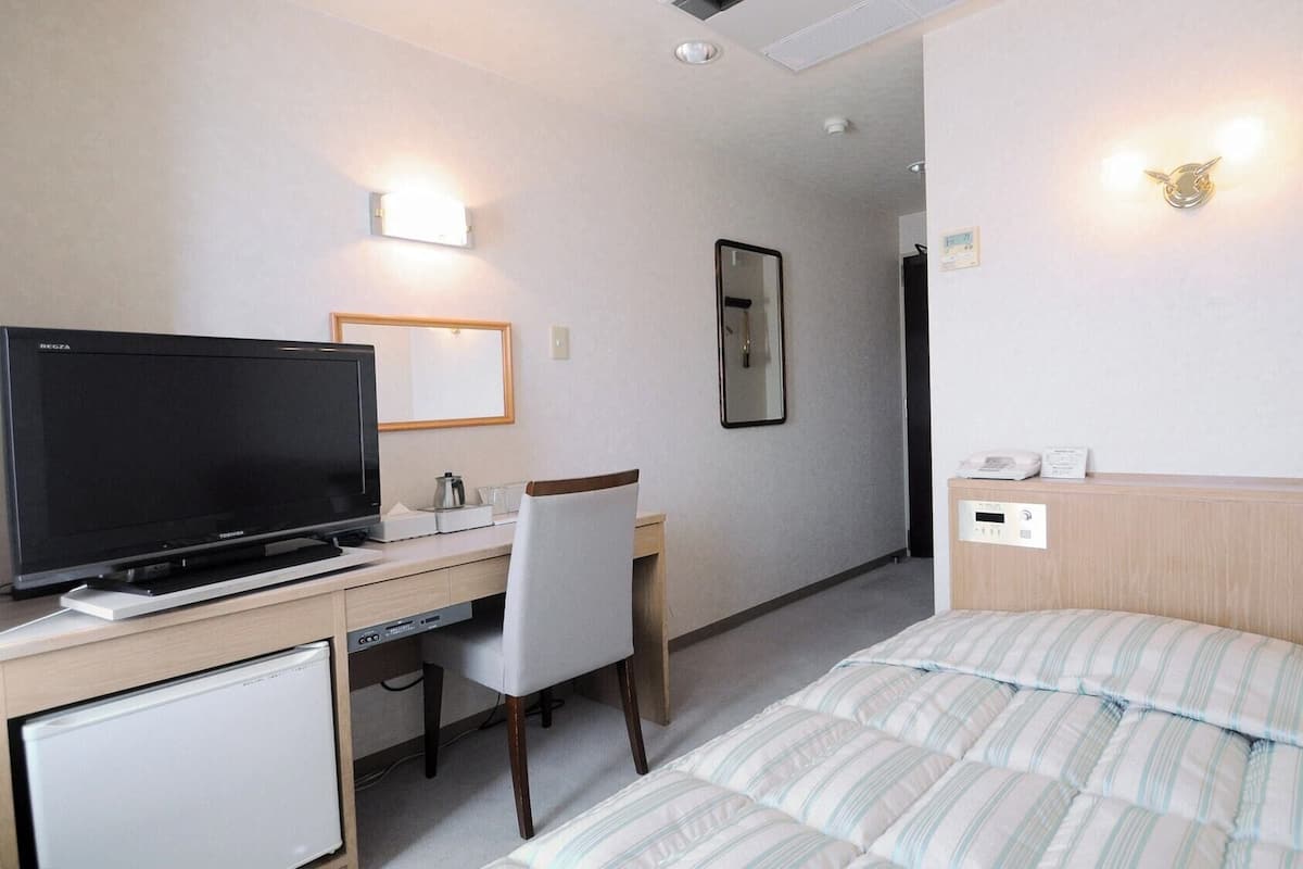 A hotel room with a bed, a desk with a television, a chair, a bedside table, and a wall-mounted light fixture.