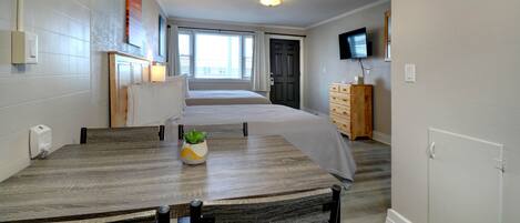 A modern hotel room with a wooden dining table, a small potted plant, a bed with a grey bedspread, a television mounted on the wall, and a window with curtains.