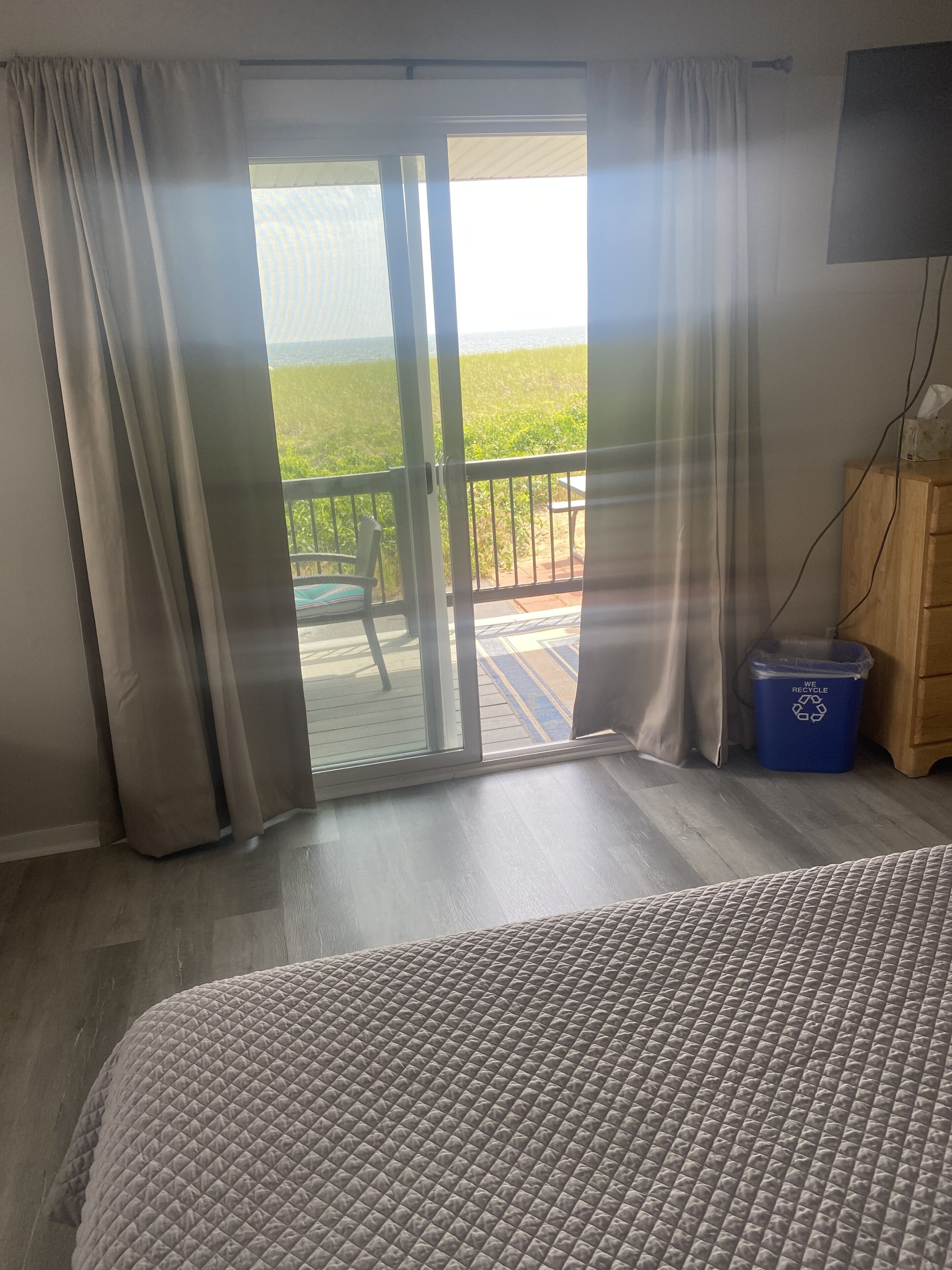 A room with a bed, a wooden cabinet, a blue recycling bin, and a balcony with a chair.