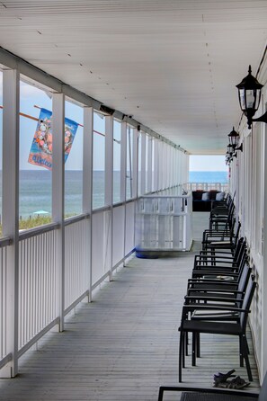 Ein überdachter Gang mit Reihen schwarzer Stühle, einer weißen Brüstung und Blick auf das Meer.