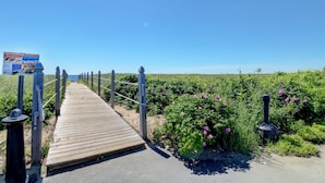 A wooden boardwalk leading through a garden with pink flowers and a clear blue sky. - On the Beach Motel (Old Orchard Beach)