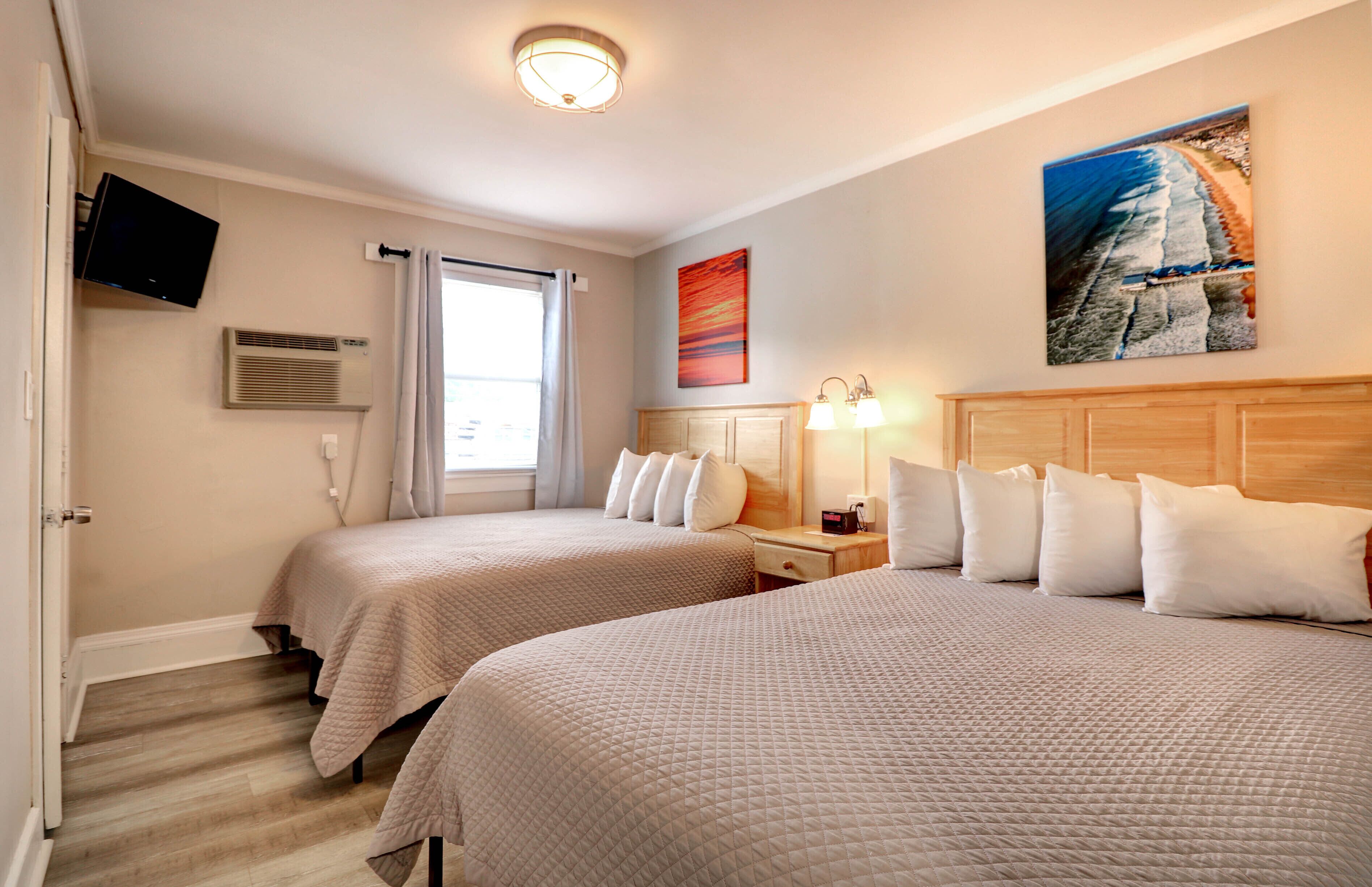 A hotel room with two beds, a television, an air conditioning unit, a painting, and a window with curtains.