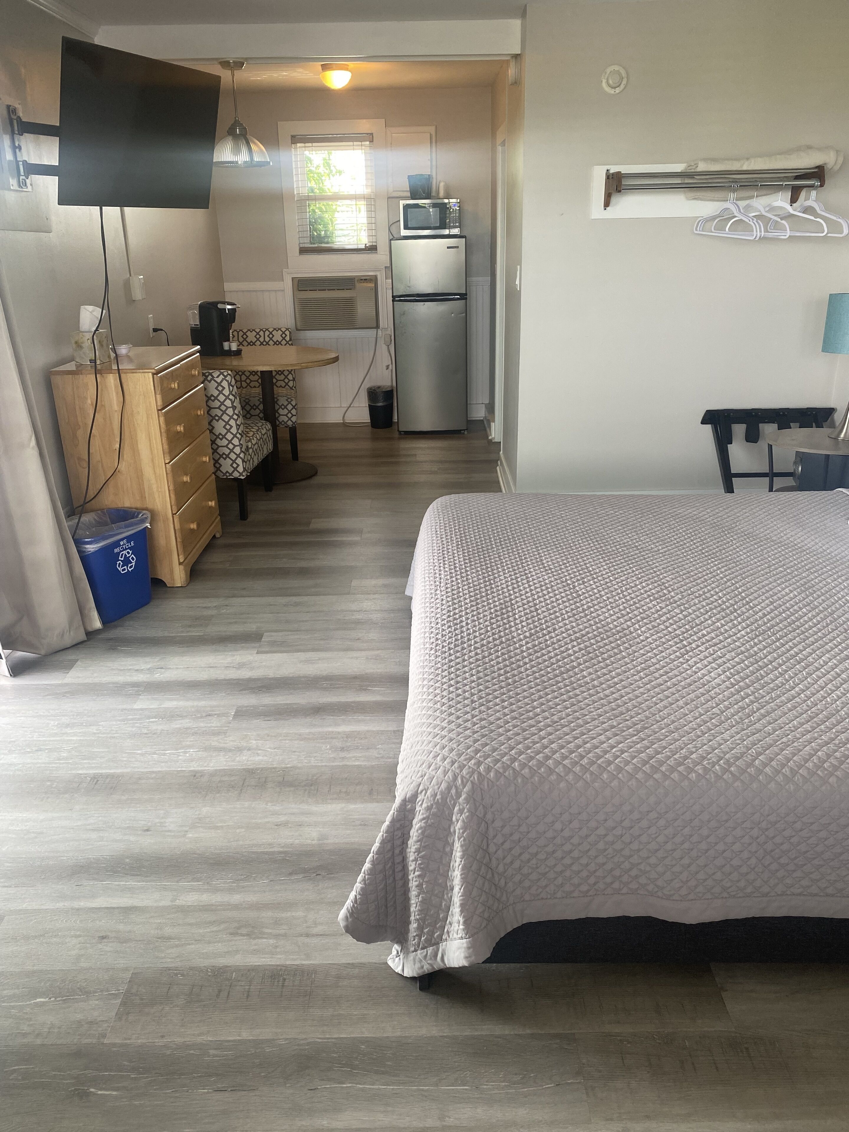 Una habitación compacta con cama, escritorio con computadora, kitchenette pequeña y televisor montado en la pared.