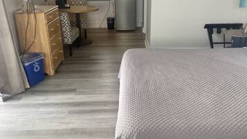 Una habitación compacta con cama, escritorio con computadora, kitchenette pequeña y televisor montado en la pared.