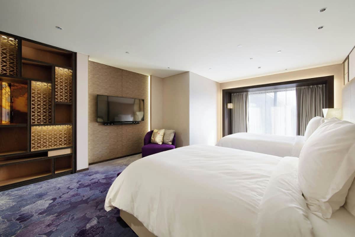 Ein Hotelzimmer mit einem großen Bett, einem an der Wand montierten Fernseher, einer Sitzecke mit einem violetten Sessel und einem Fenster mit Vorhängen.