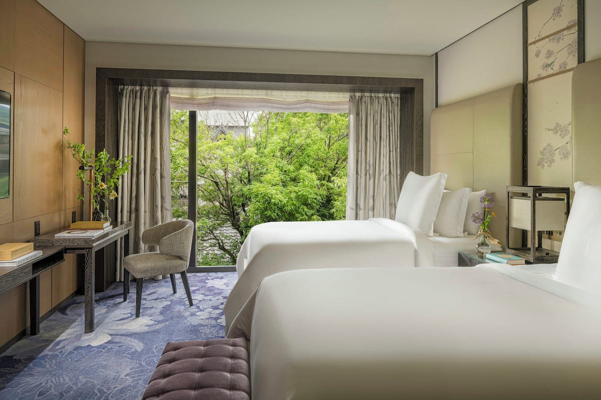 Ein Hotelzimmer mit zwei Betten, einem Schreibtisch, einem Stuhl und Blick auf Grünfläche.