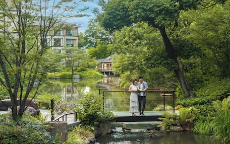 A couple standing on a bridge over a pond with lush greenery and buildings in the background.