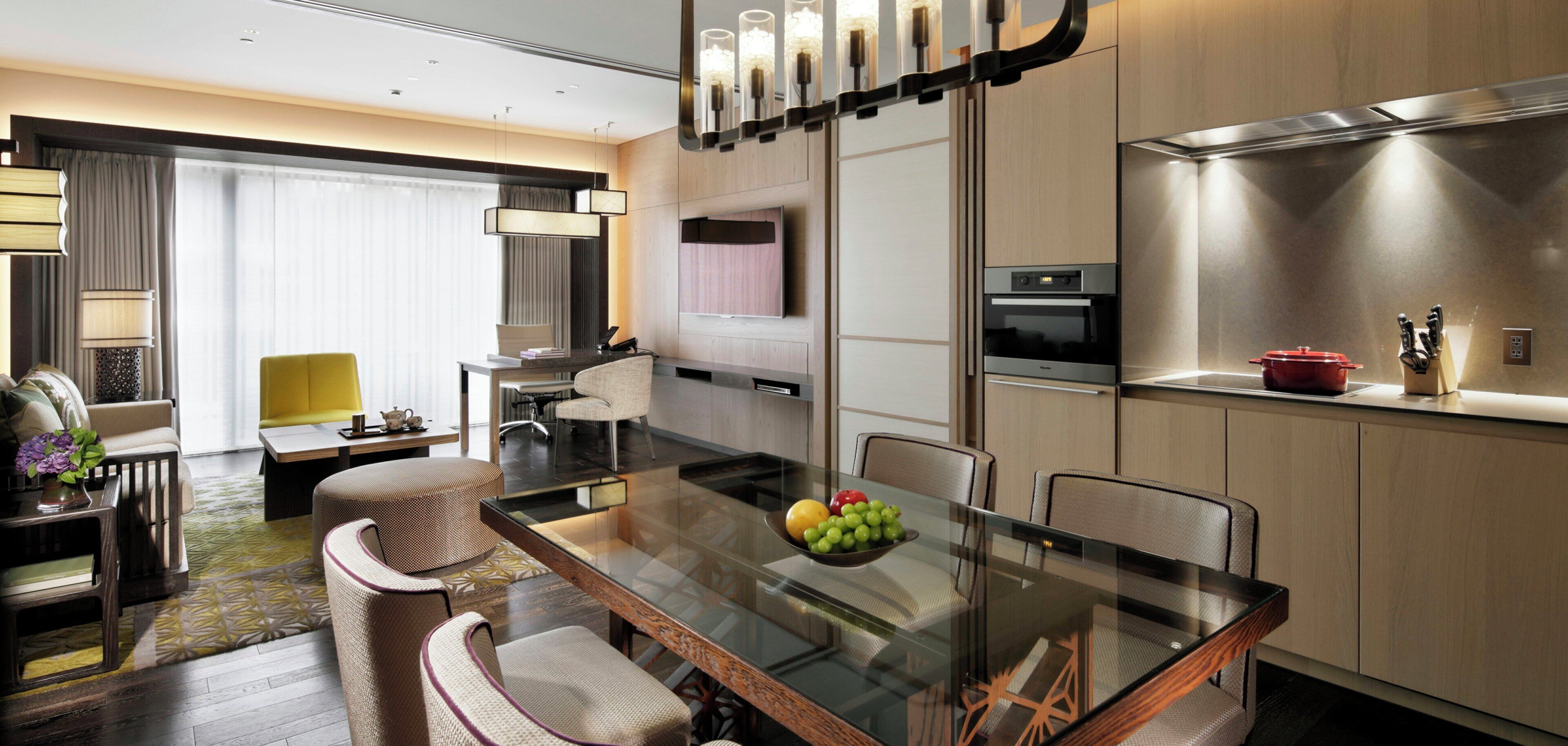 A modern hotel room with a glass dining table, a kitchen area with a red pot, and a seating area with a yellow chair.