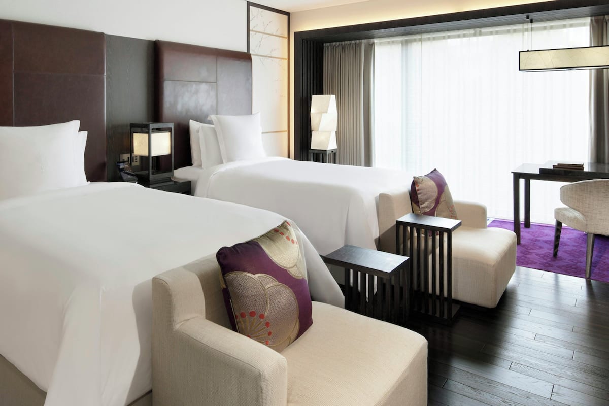 Ein Hotelzimmer mit zwei Betten, einem Schreibtisch, einem Stuhl und einem großen Fenster mit Vorhängen.