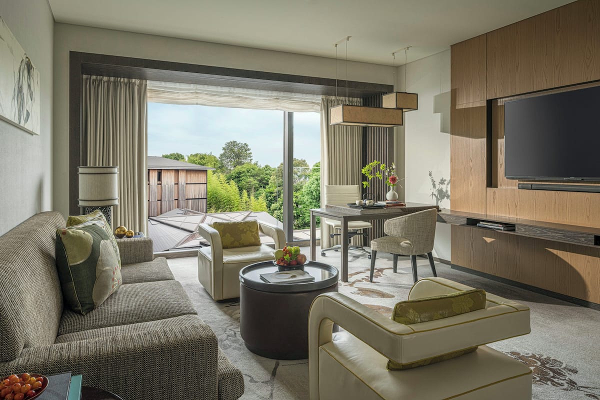Ein modernes Wohnzimmer mit einer Couch, Sesseln, einem Couchtisch und einem Flachbildfernseher. Es gibt eine Essecke mit Tisch und Stühlen sowie ein großes Fenster mit Blick auf Grünflächen.
