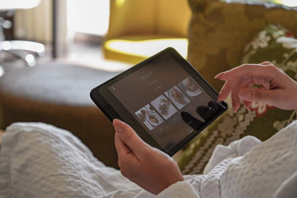 Eine Person hält ein Tablet mit einer Speisekarte in der Hand und sitzt auf einer Couch mit Kissen und Kopfstütze.