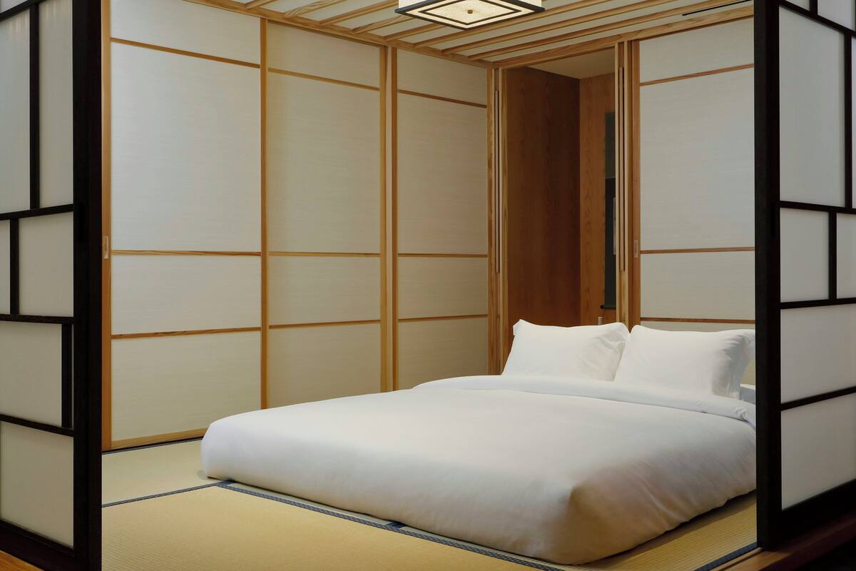 Ein traditioneller japanischer Raum mit Tatami-Bodenbelag, einem niedrigen Bett mit weißer Bettwäsche und verschiebbaren Shoji-Bildschirmen.