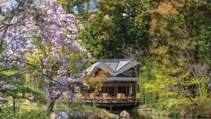 Ein traditionelles japanisches Gebäude mit einer Holzterrasse mit Blick auf einen Teich, umgeben von üppigem Grün und blühenden Bäumen.