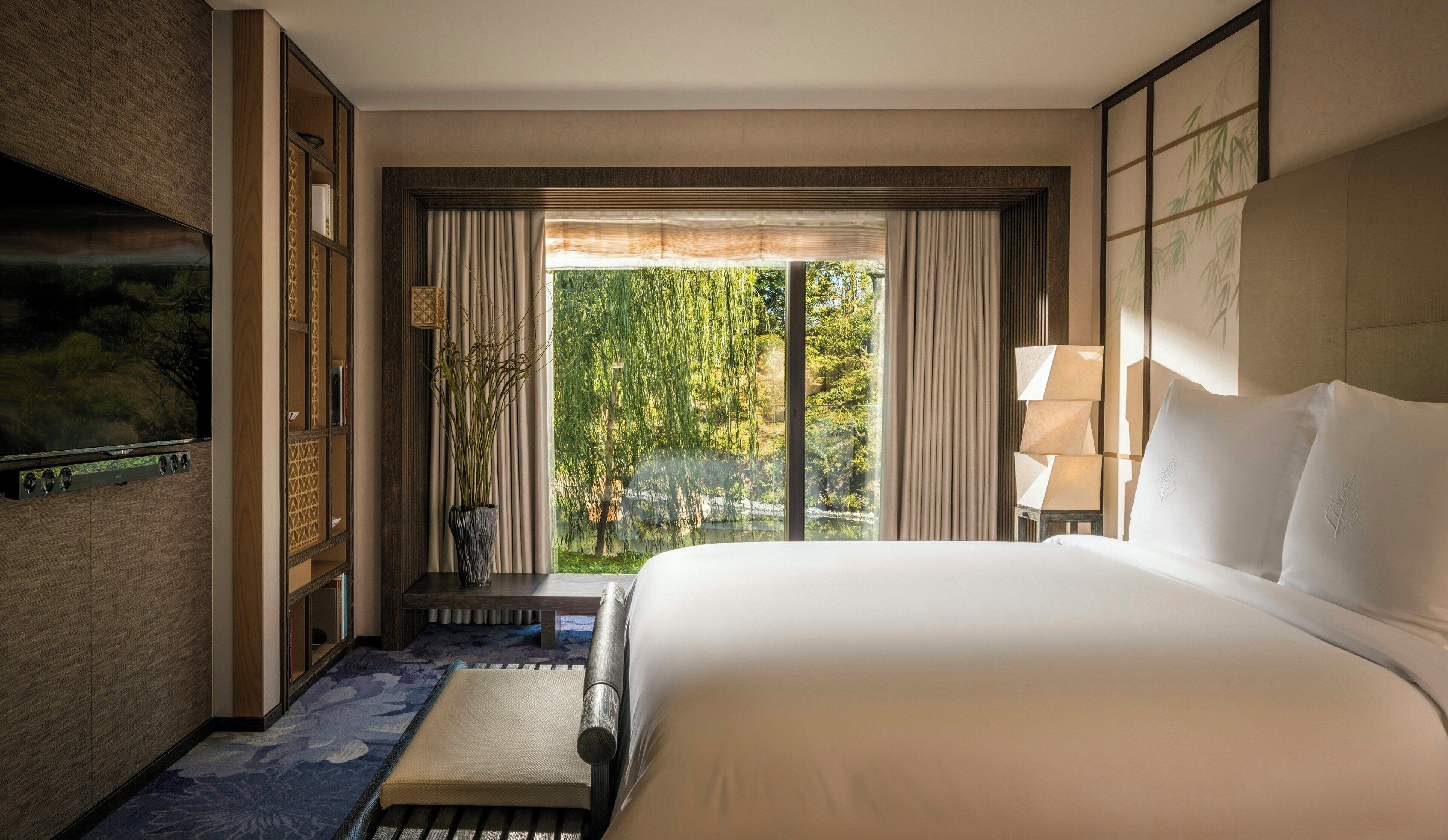 A hotel room with a large bed, a view of greenery through the window, a flat-screen TV mounted on the wall, and a bedside table with a lamp.