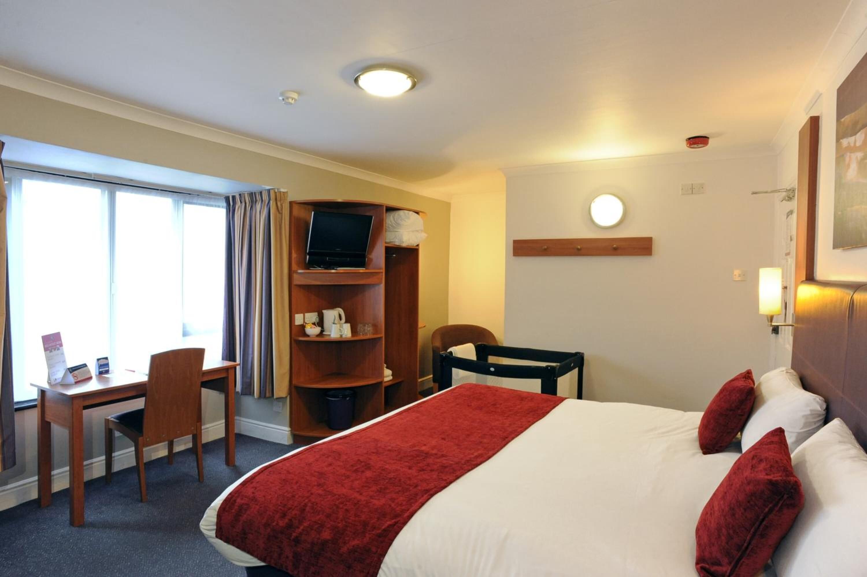 A hotel room with a bed, a desk, a chair, a TV, and a shelf with items.