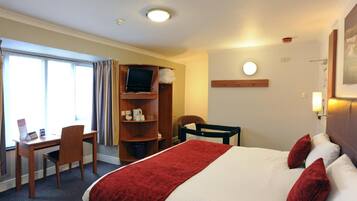 A hotel room with a bed, a desk, a chair, a TV, and a shelf with items.