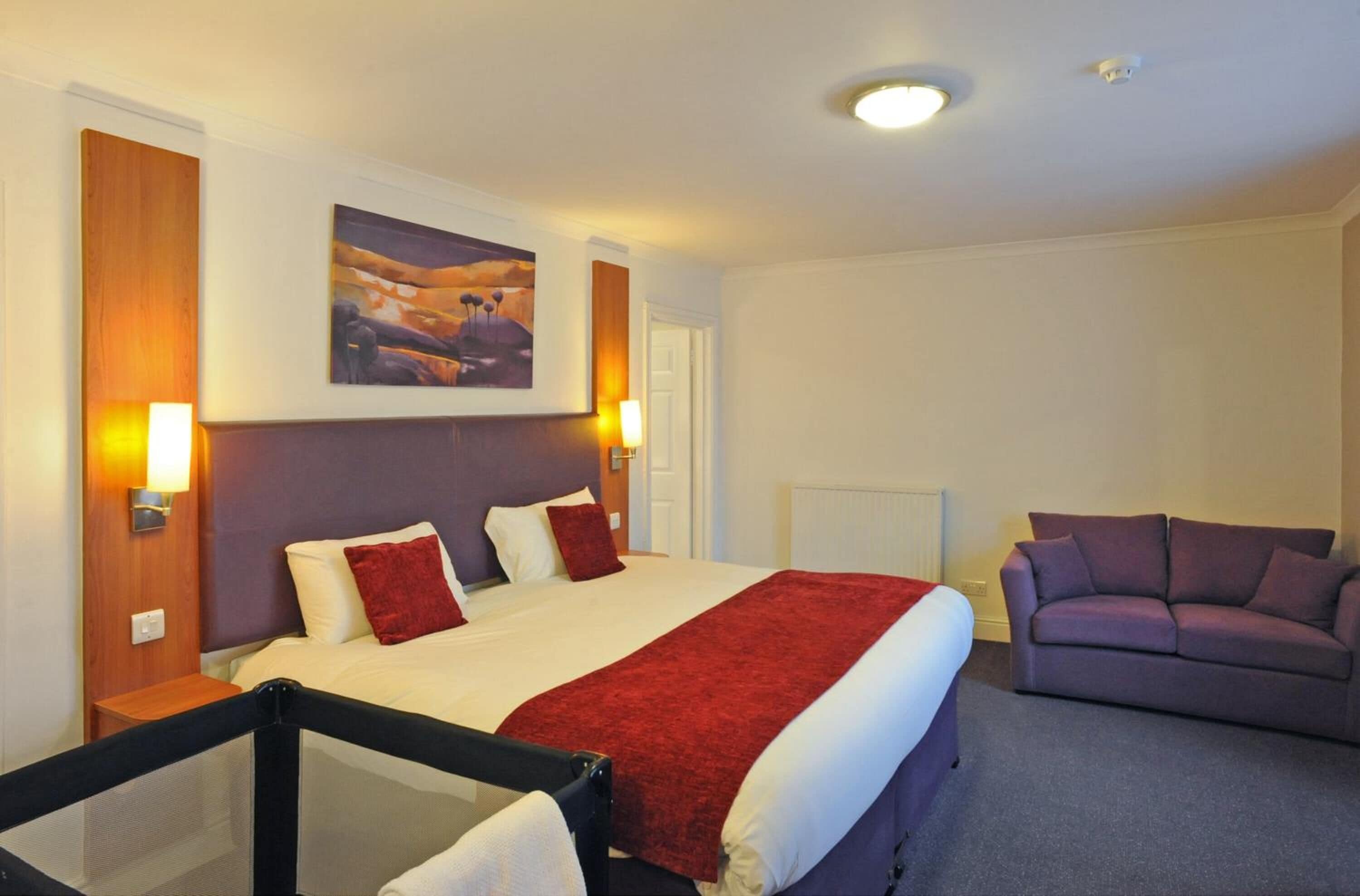 A hotel room with a bed, a red throw blanket, a purple sofa, and a painting on the wall.