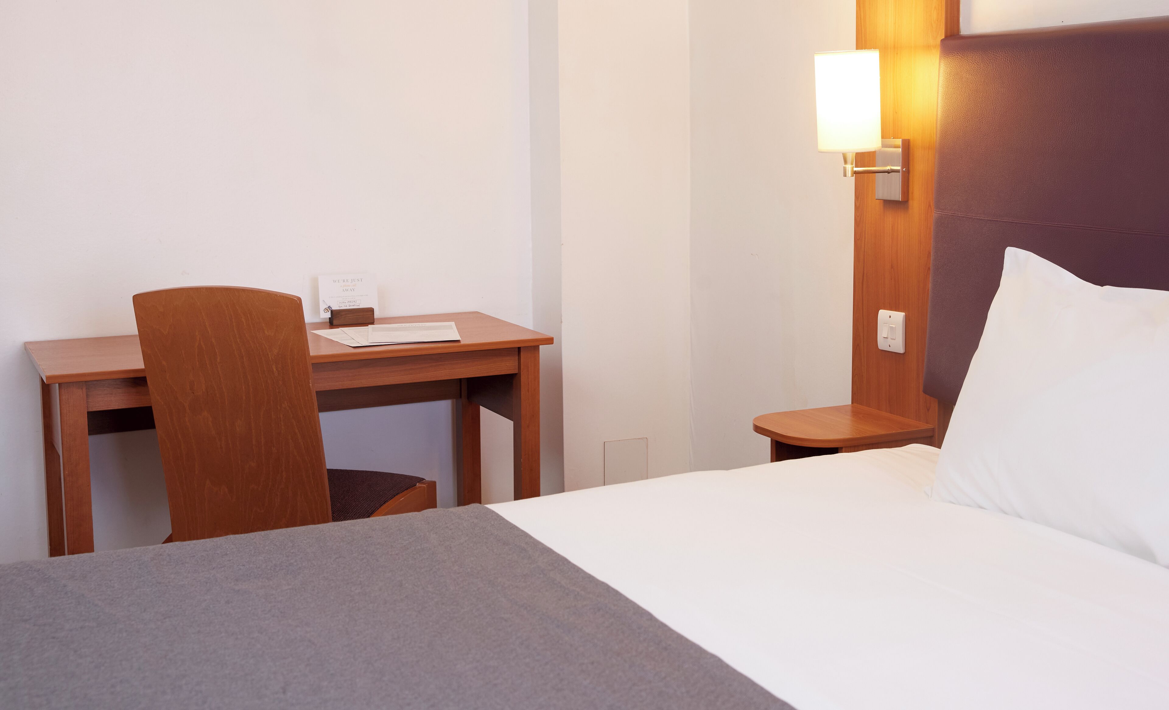 Une chambre d’hôtel avec un lit, un bureau et une chaise. Il y a un panneau sur le bureau et une lampe fixée au mur.