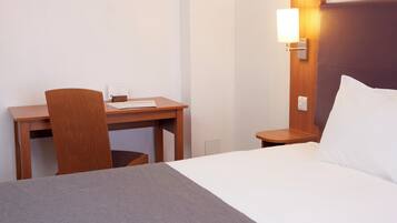 Une chambre d’hôtel avec un lit, un bureau et une chaise. Il y a un panneau sur le bureau et une lampe fixée au mur.