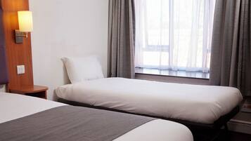 A hotel room with two single beds, a bedside table with a lamp, a wooden headboard, and a window with curtains.