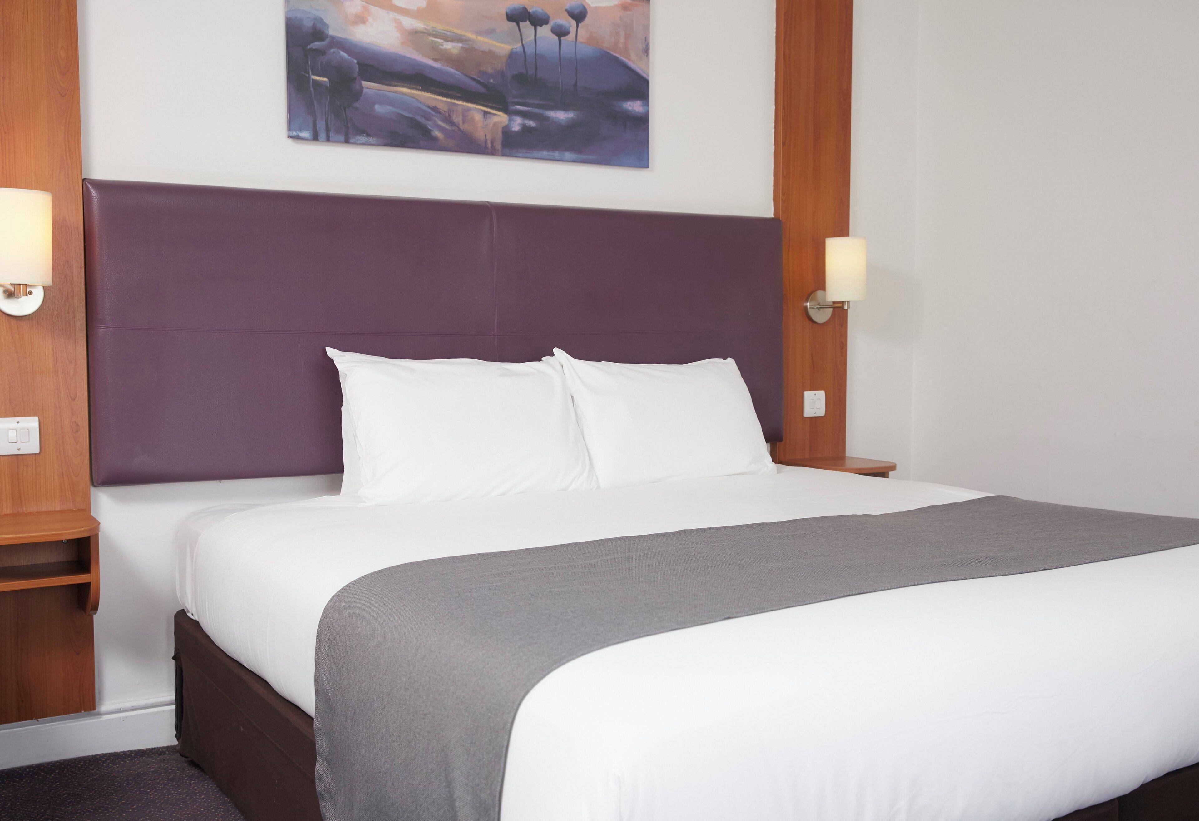 A neatly made bed with white linens and a gray throw blanket, a purple headboard, and two wall-mounted lamps on either side.