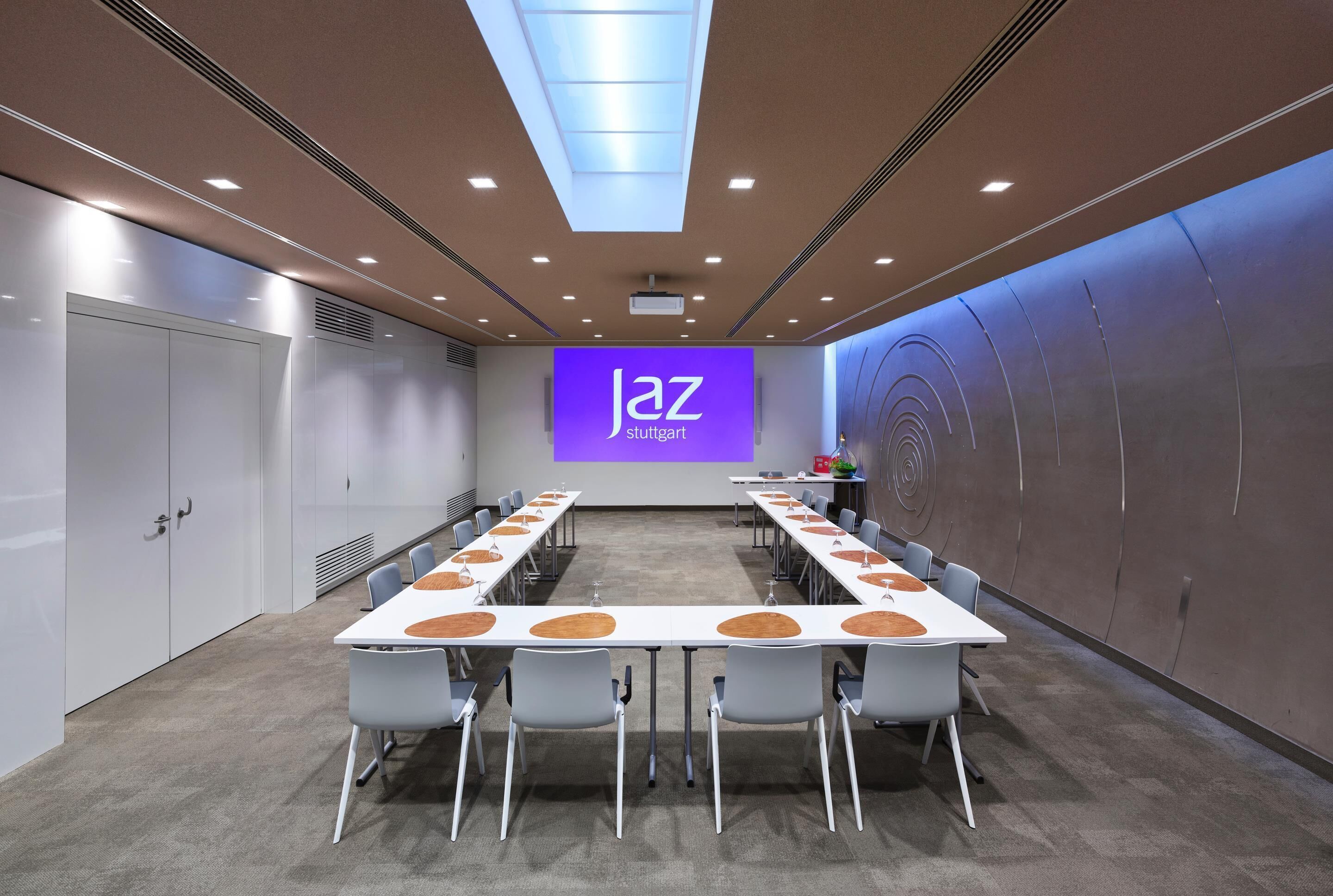 Un salón de conferencias con una larga mesa, sillas, una pantalla de proyector y una pared con un logotipo.