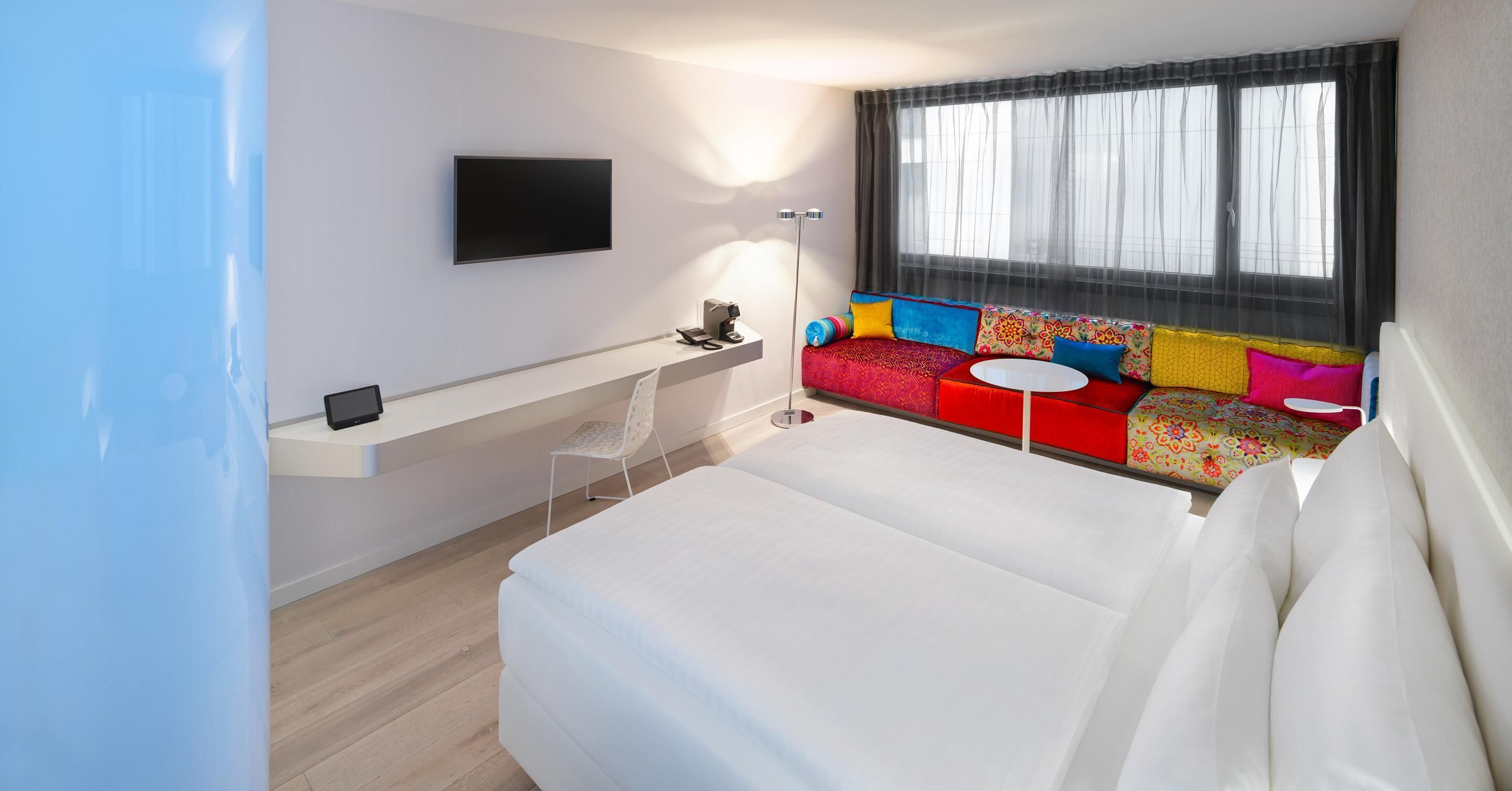 A modern hotel room with a large bed, a desk with a flat-screen TV, a sofa with colorful cushions, and a large window with sheer curtains.
