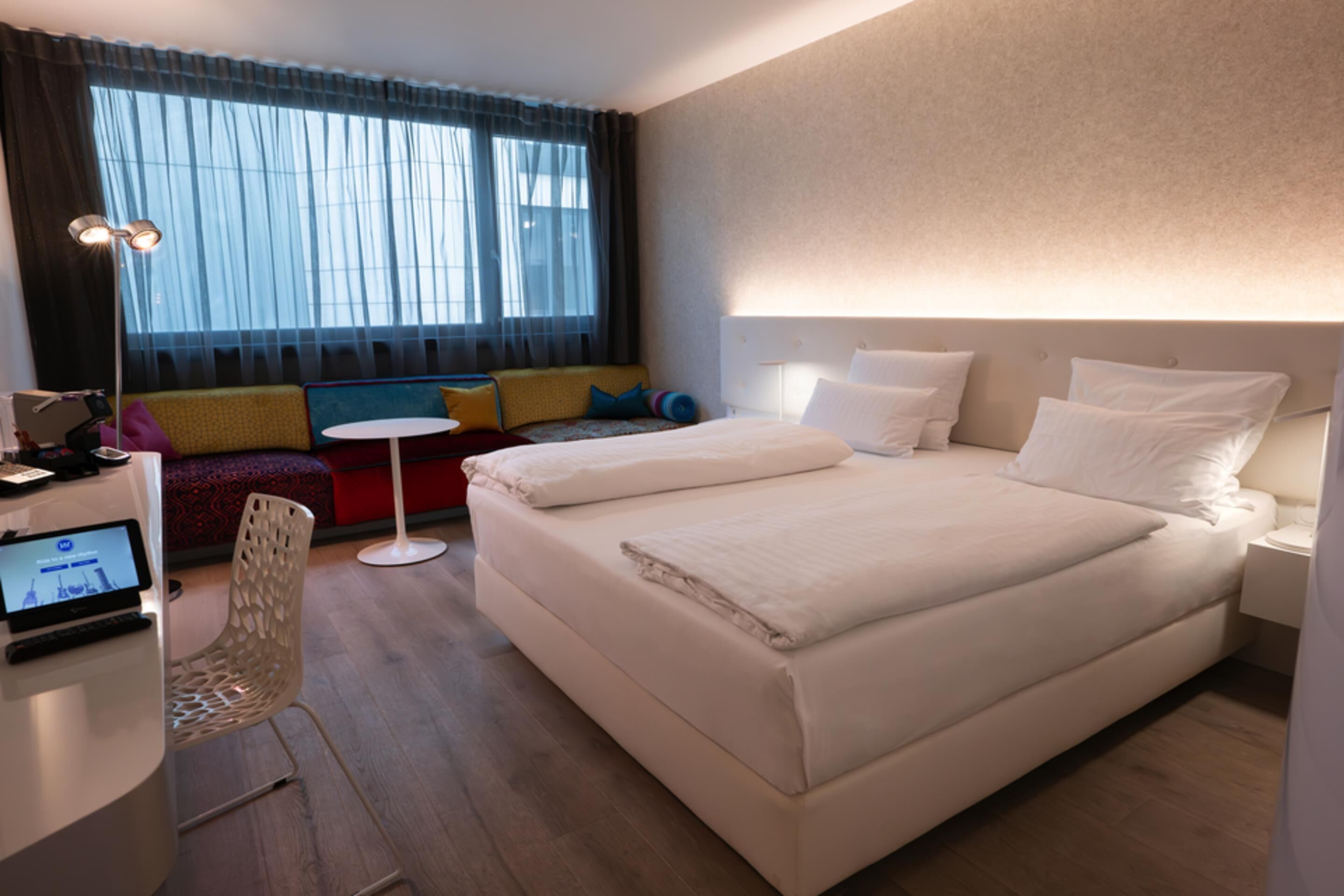 Ein Hotelzimmer mit einem Bett, einem Schreibtisch mit Laptop, einem Sessel und einer Couch, sowie einem Fenster mit Vorhängen.