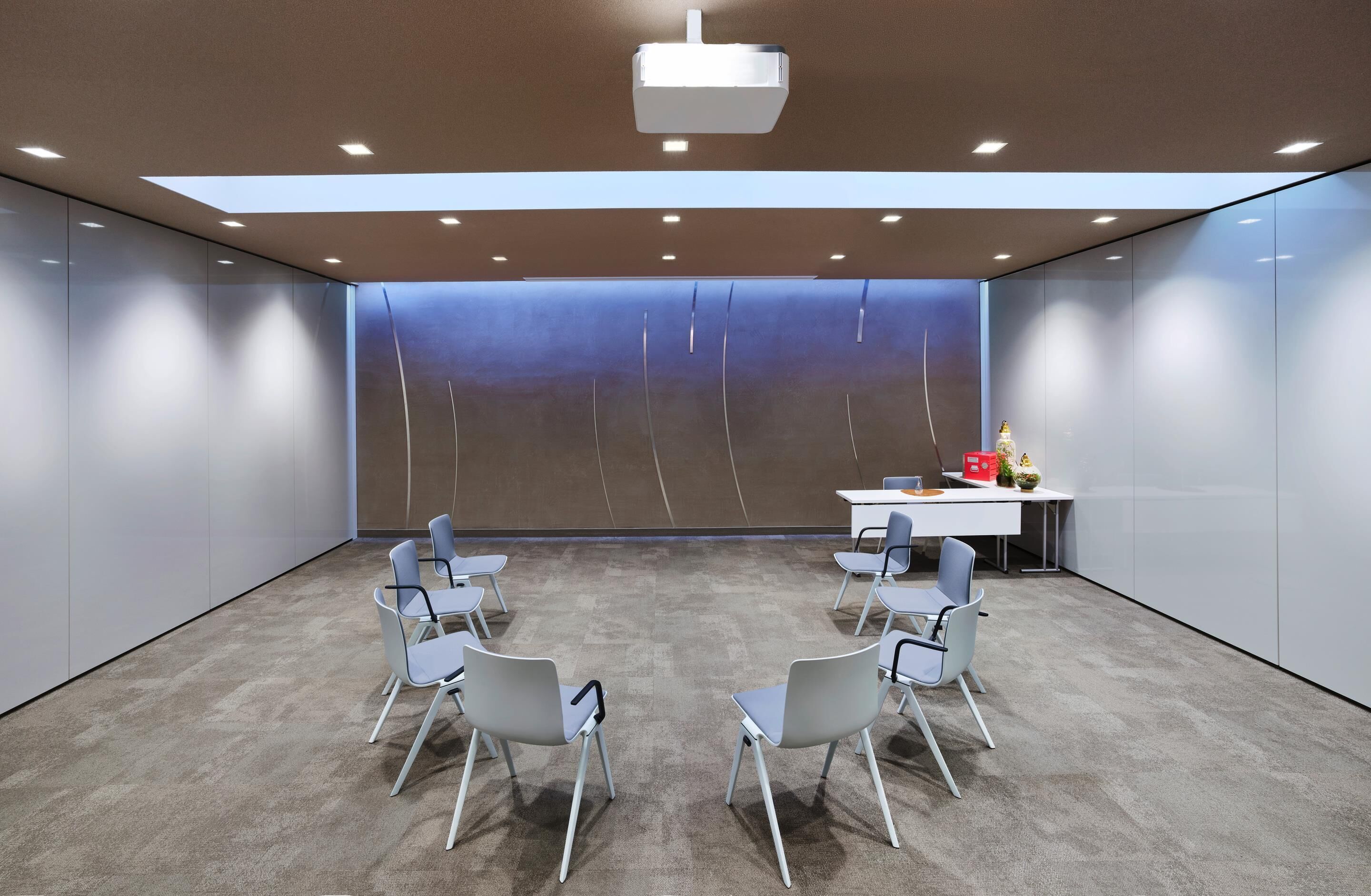 A modern meeting room with a high ceiling, recessed lighting, a central table with chairs, and a large screen.