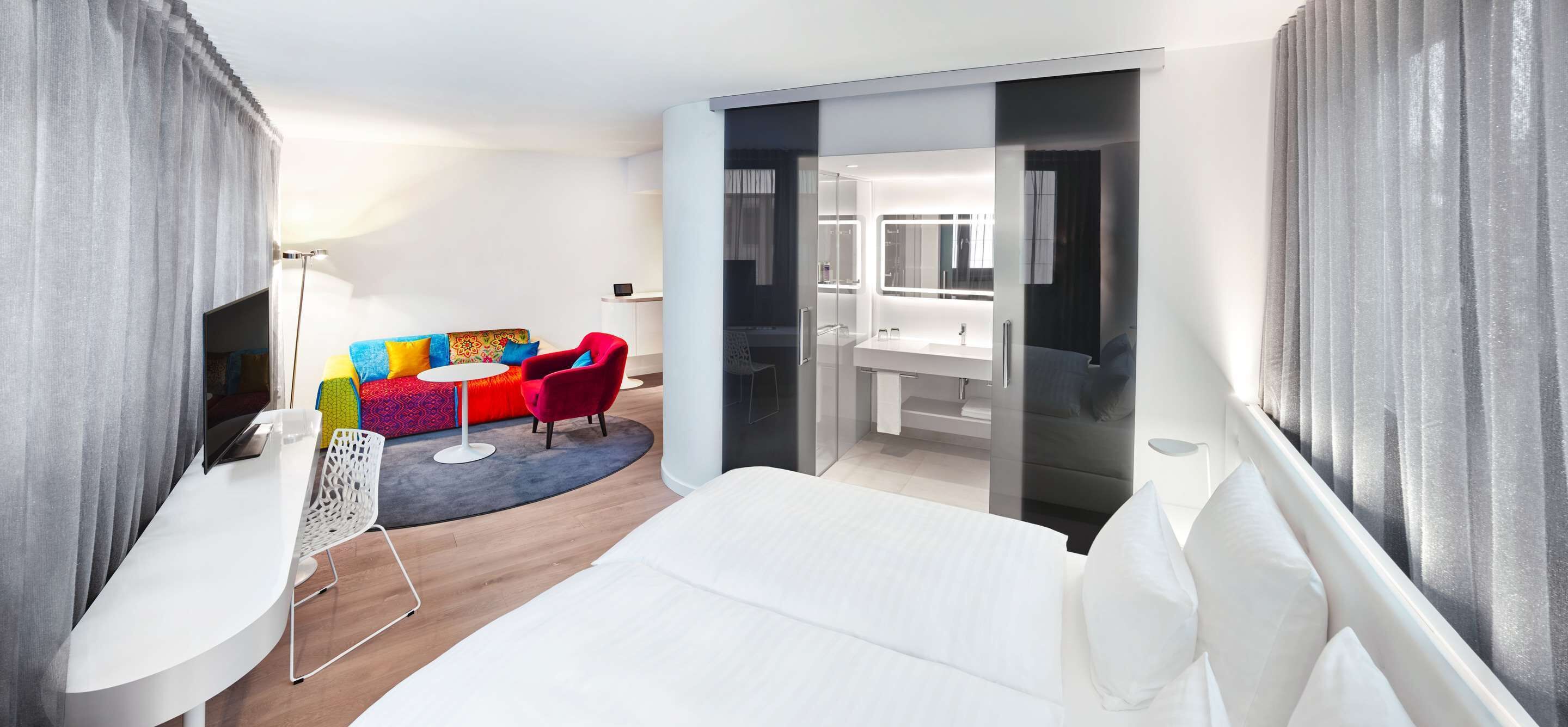A modern hotel room with a bed, a small seating area with colorful cushions, a desk, and a bathroom with a mirror and sink.