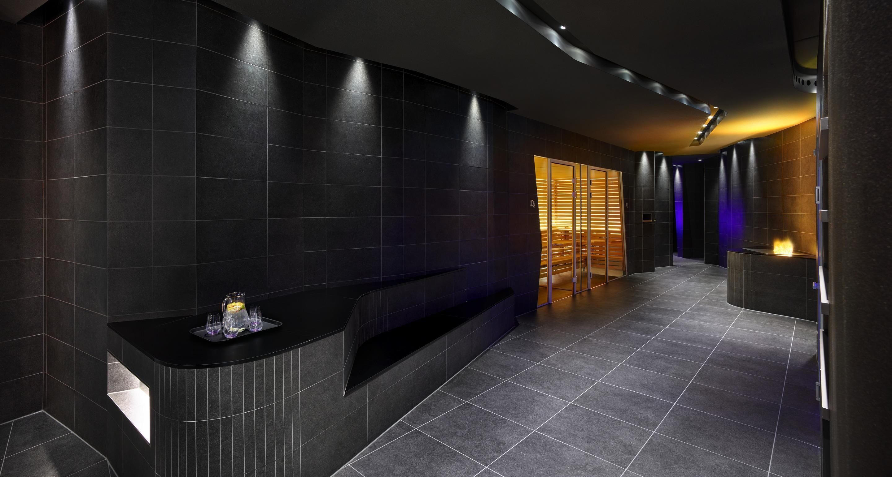A modern bathroom with dark tiled walls, a curved counter, and a glass enclosure with a warm light.