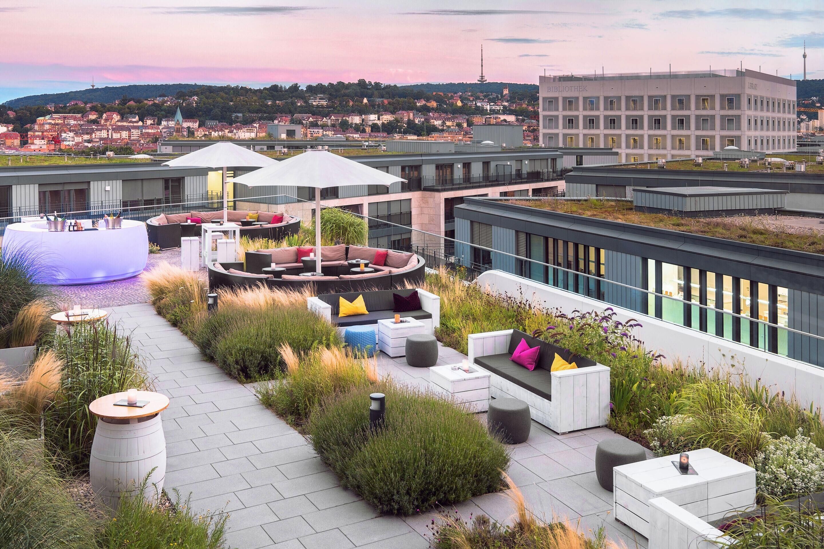 Una terraza en la azotea con mobiliario exterior, vistas a la ciudad y un edificio moderno al fondo.
